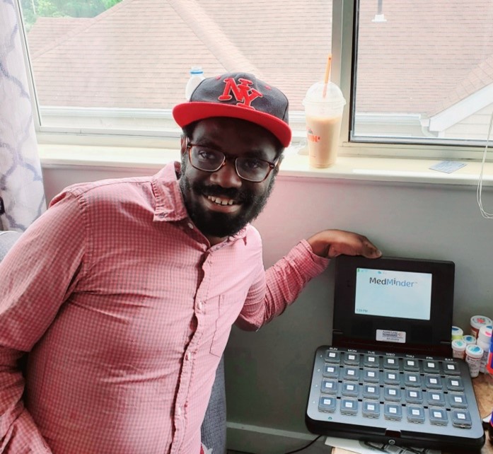 A man with a hat posing with a smile next to his MedMinder device