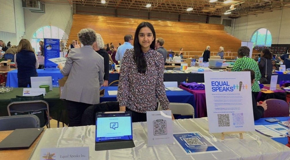 Ayana Singh stands behind an exhibitor table displaying the ConnectAAC app on a laptop and a QR code sign to try the app, with an Equal Speaks poster beside her at an event with other exhibitor tables and attendees in the background.