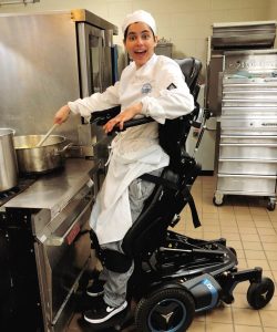 Young woman in cook's outfit, utilizing a standing wheelchair, as she stirs a pot in an industrial kitchen. She has a big smile on her face.